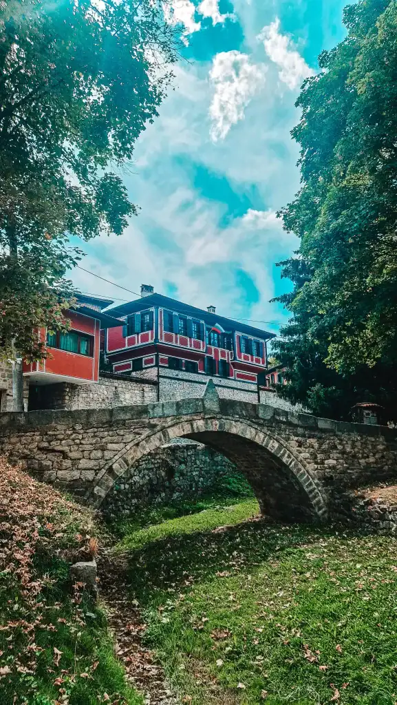 Stone arch bridge over a dry stream, leading to a traditional red and black Bulgarian Revival-style house surrounded by lush trees under a partly cloudy sky. This historic scene captures the charm of Koprivshtitsa’s preserved architecture.