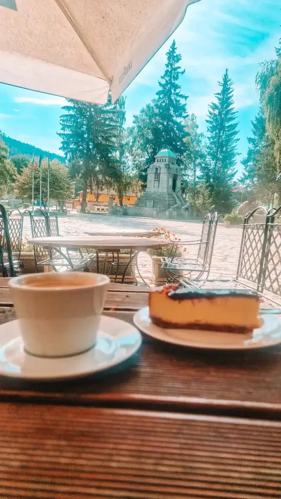 Cup of coffee and a slice of cheesecake on a café table with a view of a cobblestone square and a stone monument in the background. Enjoying local cafés is one of the most relaxing things to do in Koprivshtitsa.