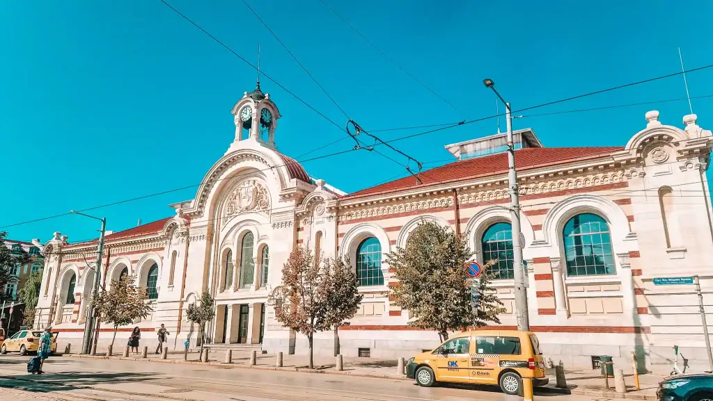 A grand neoclassical building with a red-tiled roof and clock tower stands along a tram-lined street under a clear blue sky. The architectural detail reflects Bulgaria's cultural heritage.