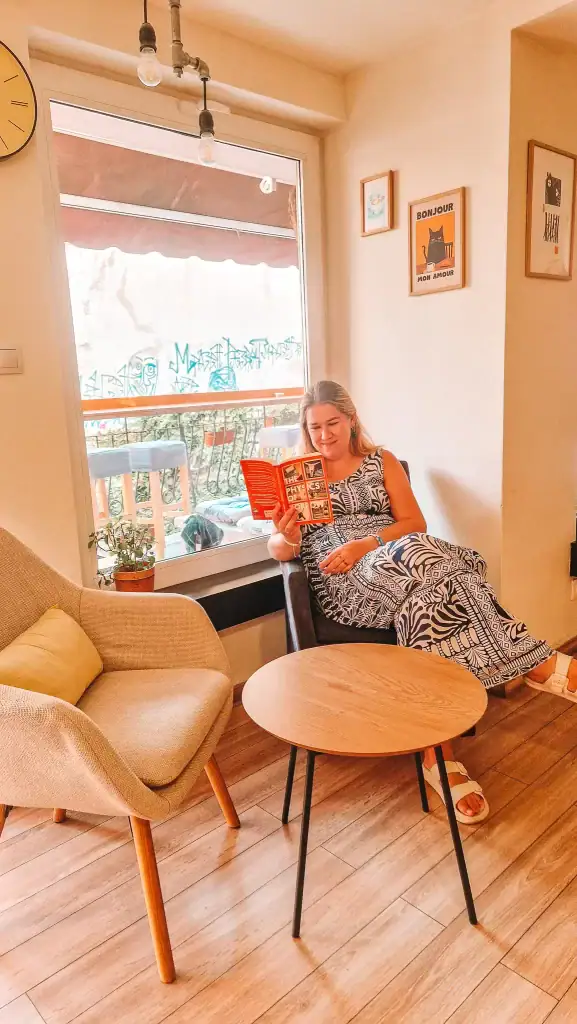 Em relaxes in a cosy corner of a café, smiling while reading a bright red copy of The Physics of Sorrow by Georgi Gospodinov. The stylish indoor setting and Bulgarian literature in hand highlight one of the most thoughtful books about Bulgaria.