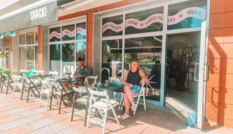 Em sits outside a café called SNACK! Specialty Coffee, smiling beside a laptop and iced drink while another customer checks their phone nearby. The sunlit patio is set with white folding chairs and small round tables.