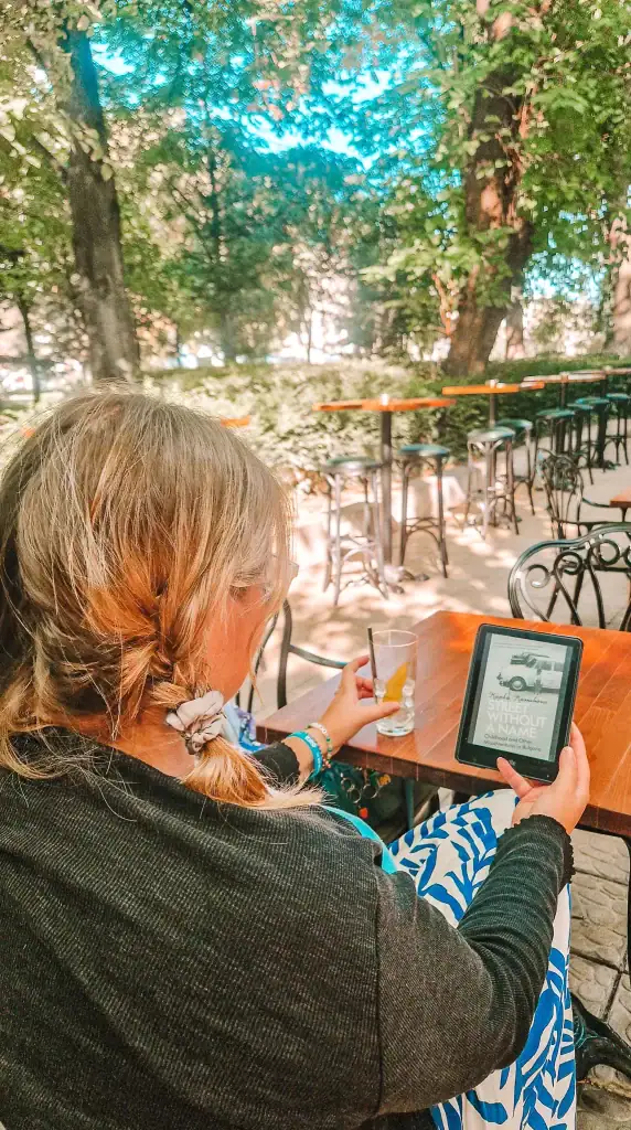 Em sits at an outdoor café under leafy trees, holding a Kindle that displays Street Without a Name by Kapka Kassabova, with a half-finished citrus drink on the table. A quiet moment with one of the most personal books about Bulgaria.