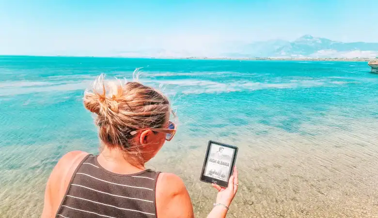 Em stands in shallow turquoise waters holding an e-reader that displays the cover of High Albania by Edith Durham. The clear lake reflects distant mountains beneath a cloudless sky, tying literature to the Albanian landscape.