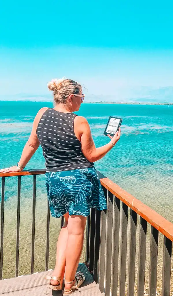 Em leans against a wooden railing while reading High Albania on her e-reader. She overlooks a panoramic view of a bright blue lake with Albanian mountains fading into the background, highlighting her exploration of books about Albania in the places they describe.