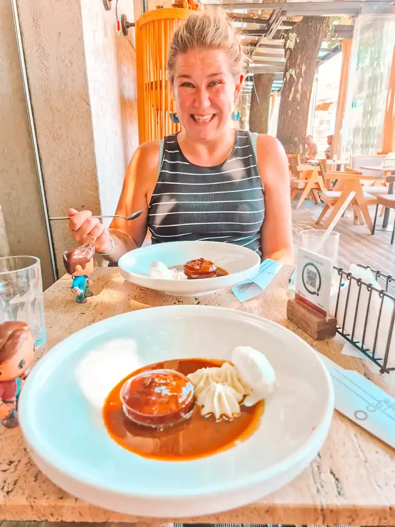 Em grins while seated at a shaded outdoor café in Shkoder, about to enjoy a dessert of sticky toffee pudding, served with whipped cream and a scoop of ice cream on a white plate.