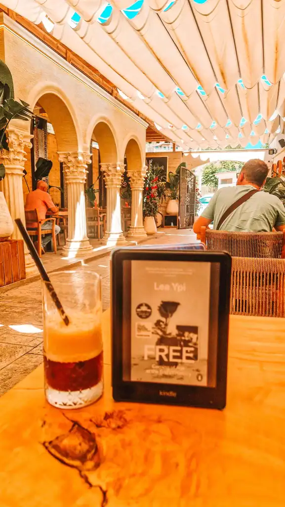 An e-reader showing the cover of Free by Lea Ypi rests on a sunlit café table beside a half-finished iced coffee. Ornate arches and leafy plants surround the patio, creating a calm space to explore books about Albania’s communist past.