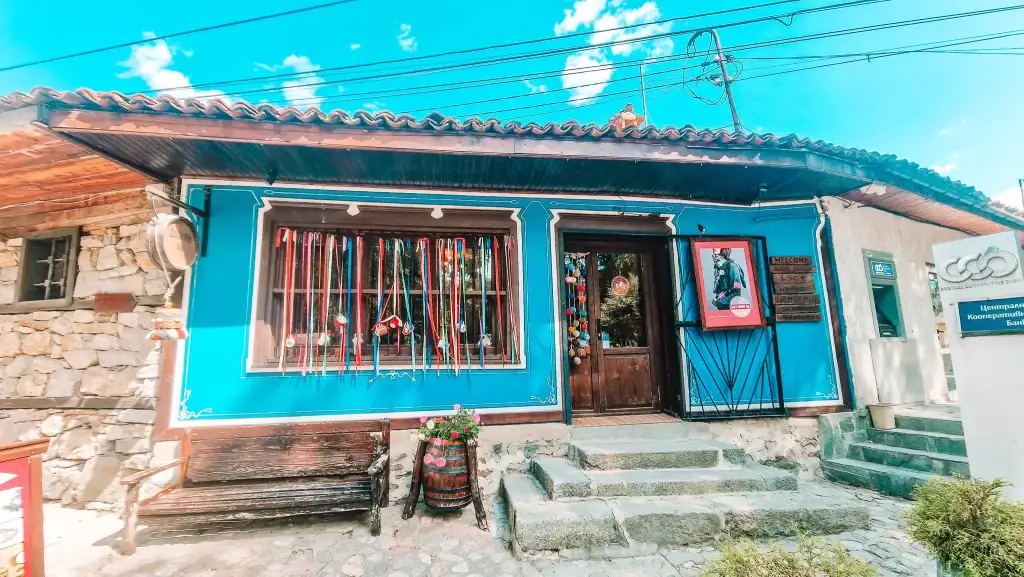 Charming blue souvenir shop in Koprivshtitsa decorated with colourful ribbons and handmade crafts in the window, next to a local bank branch. Shopping for traditional Bulgarian gifts is one of the cosy things to do in Koprivshtitsa.