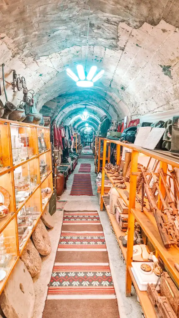 Tunnel-like museum corridor filled with Albanian military and domestic artefacts, from rotary phones to uniforms and kitchenware. Rows of red-patterned rugs lead the eye through this immersive historical exhibit.