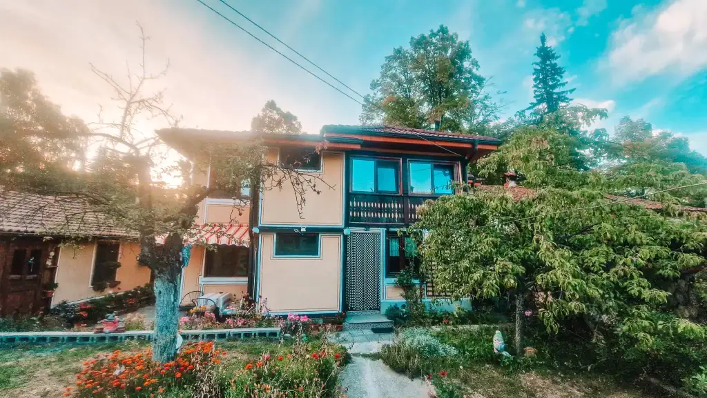 Cosy yellow house with a red-tiled roof, colourful garden, and trees framing the entrance at sunset. Staying in a local guesthouse is a peaceful way to experience life in Koprivshtitsa.