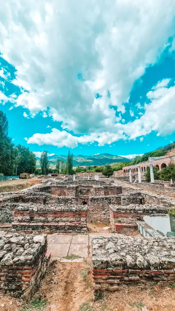 Low stone walls and partially excavated ancient structures at Heraclea, set against a mountainous Macedonian backdrop under a dramatic sky.