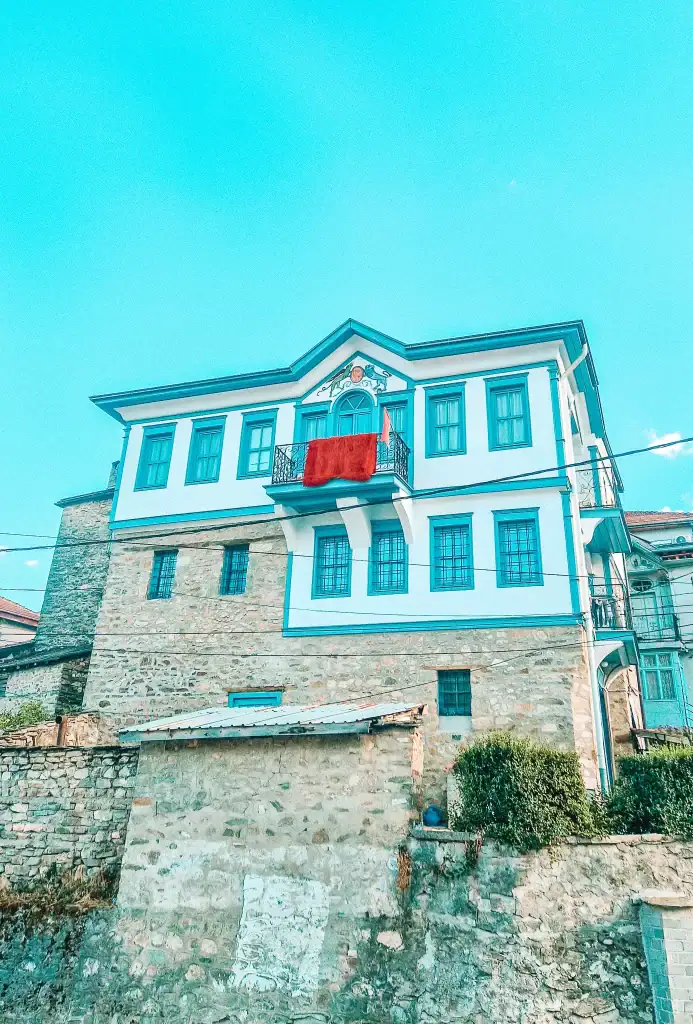 A traditional Ottoman-style house in Krusevo with blue trim and stone base, featuring a small balcony with a red woven cloth draped over the railing.