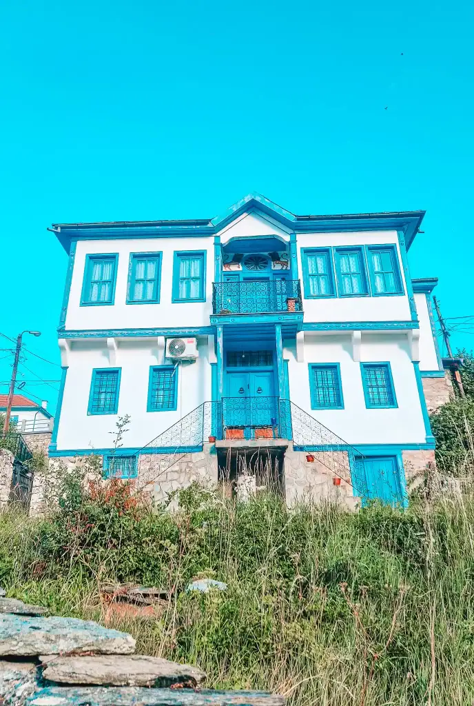 Restored white heritage house with vivid blue doors, windows, and balcony railings, surrounded by tall grass and shrubs, highlighting the town’s historic architecture.