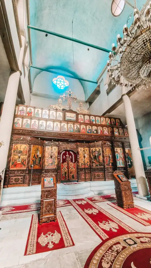 Ornate interior of an Orthodox church in Krusevo featuring a richly carved wooden iconostasis with vibrant religious icons, red carpets, and a grand chandelier hanging from a vaulted ceiling.