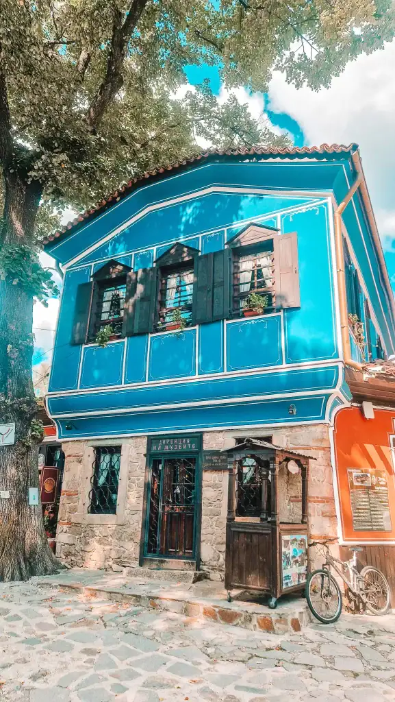 Bright blue heritage building with stone ground floor and a bicycle parked outside, surrounded by trees and cobbled streets. This museum complex is one of the many cultural stops when exploring things to do in Koprivshtitsa.