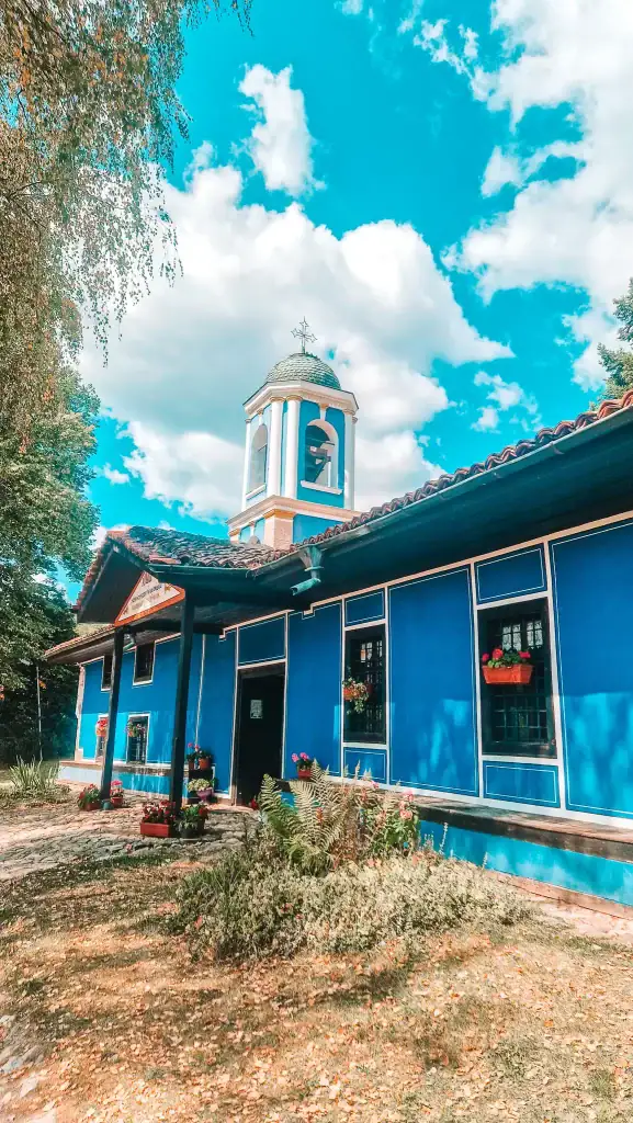 Blue-painted church with white bell tower and cross, surrounded by trees and fallen leaves, under a bright blue sky. Visiting the historic Church of the Assumption is a must for those interested in local spiritual heritage.