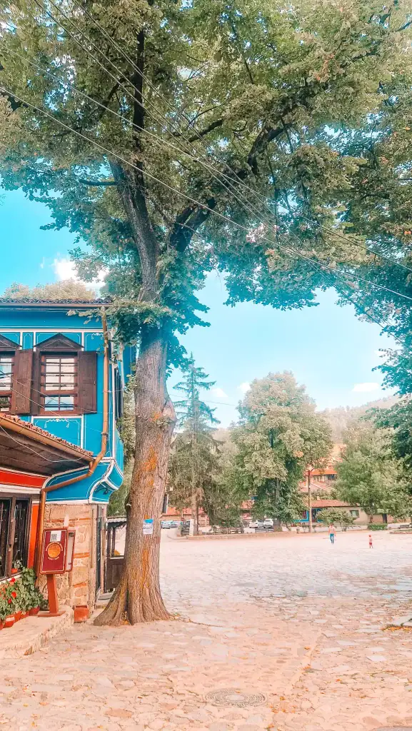 Cobblestone town square in Koprivshtitsa shaded by tall trees, with a glimpse of a vivid blue heritage building and a few people walking in the distance. Wandering through the historic centre is one of the simplest pleasures here.