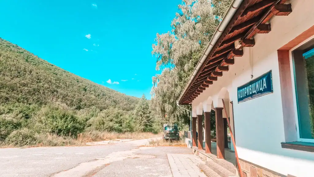 View of the Koprivshtitsa train station with a blue sign in Cyrillic, surrounded by lush hills and rustic charm. Taking the scenic train route is a relaxing way to arrive for a day of exploring things to do in Koprivshtitsa.
