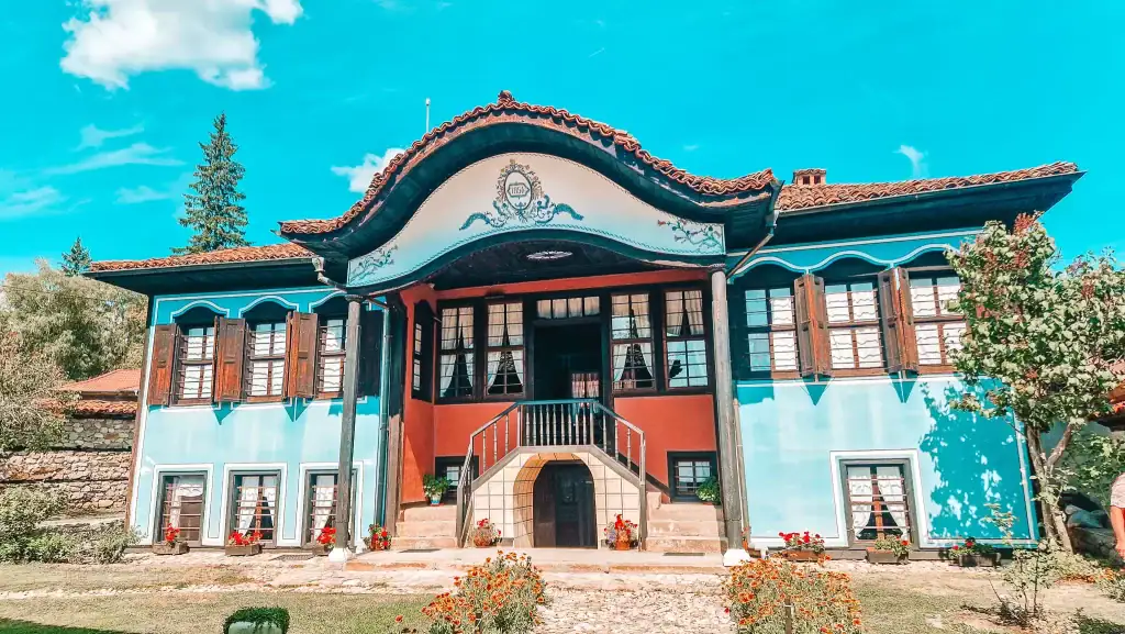 Front view of the Lyutov House with its curved roofline, red and blue façade, wooden shutters, and bright flowerpots under a clear sky. This well-preserved mansion is one of the key cultural things to do in Koprivshtitsa.