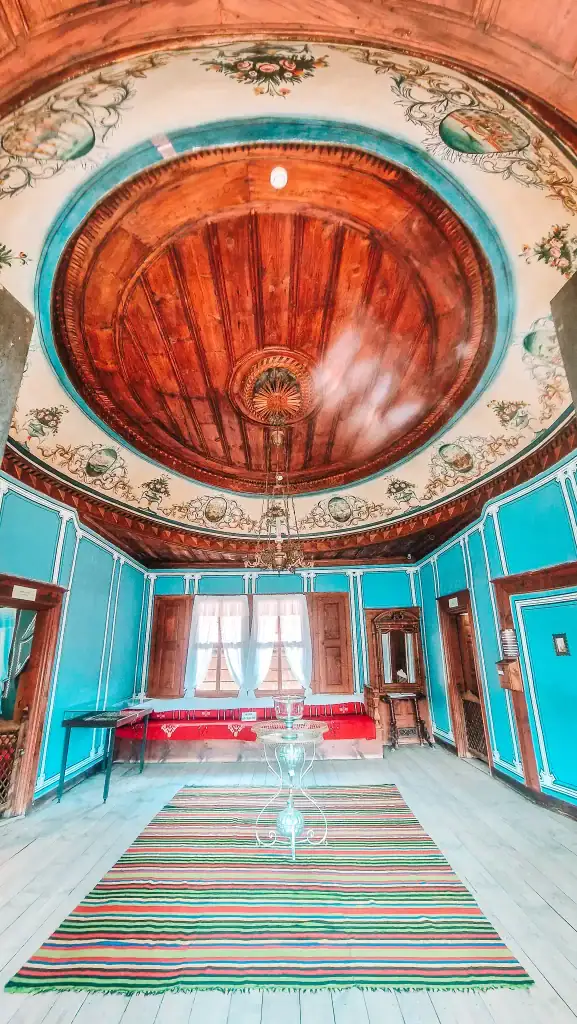 Interior of a historic house museum featuring turquoise walls, a carved wooden ceiling dome, floral frescoes, and striped traditional rugs. Visiting these restored homes offers insight into Bulgaria’s Revival period.