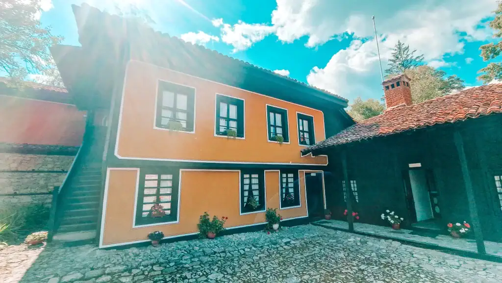 Courtyard view of a traditional orange and black two-storey house with cobbled ground, flower pots, and a rustic tiled roof. The architectural style showcases the preserved beauty of this historic town.