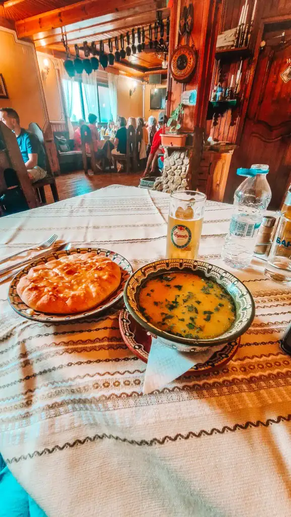 Traditional Bulgarian meal served in a rustic tavern, featuring a bowl of soup, fluffy flatbread, and a glass of Zagorka beer on an embroidered tablecloth. Tasting local cuisine is a must on any list of things to do in Koprivshtitsa.