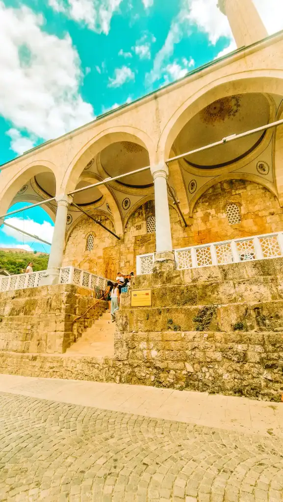 Stone exterior of the Sinan Pasha Mosque in Prizren, Kosovo, with a wide staircase, elegant arches, and visitors entering beneath its intricately painted ceilings.