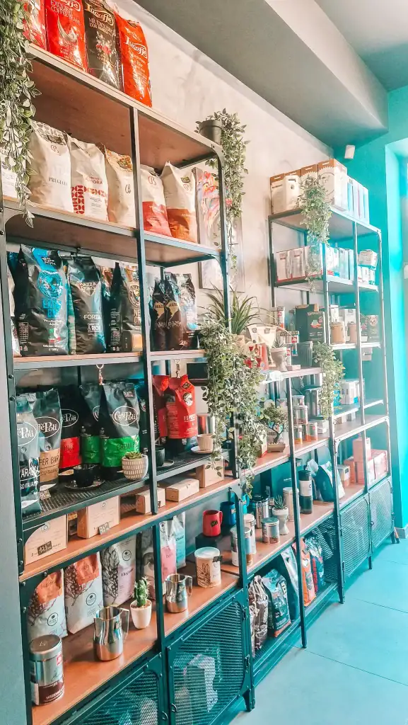 Shelves stocked with various coffee beans, equipment, and accessories in a well-lit shop corner. Decorative green plants weave through the shelves, giving a curated feel to this café retail space.