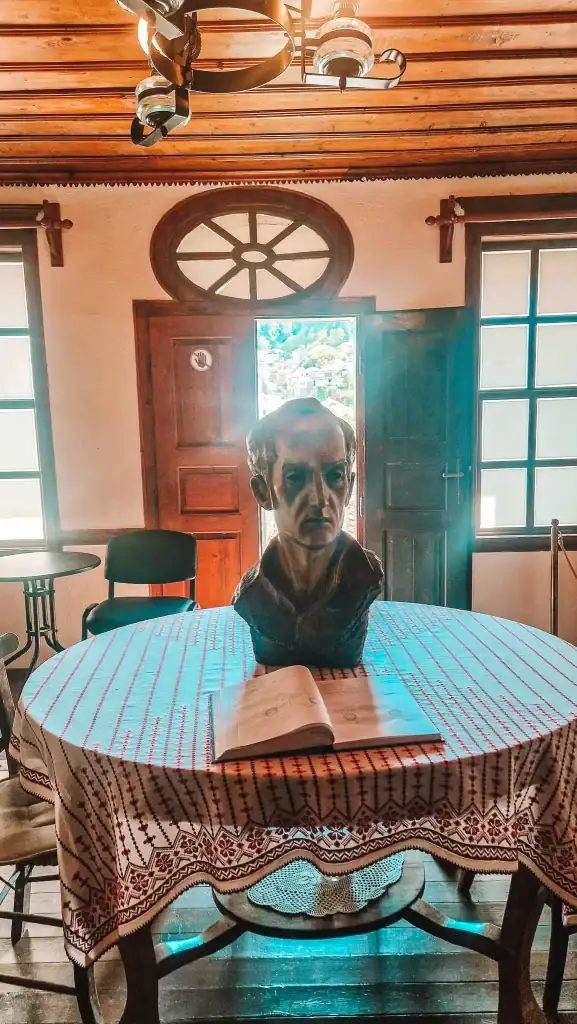 Bronze bust of a man placed on a round table covered with traditional embroidered cloth inside a heritage home, with sunlight streaming through wooden windows and a scenic view outside.