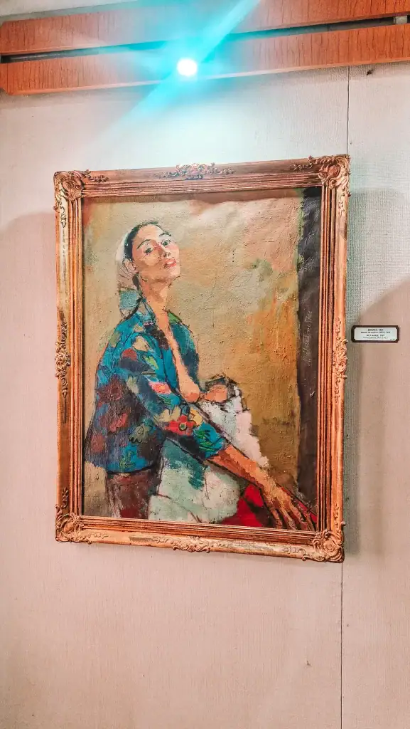 Framed oil painting of a woman with a confident pose, wearing a blue floral blouse and headscarf, displayed under a spotlight in an art gallery.