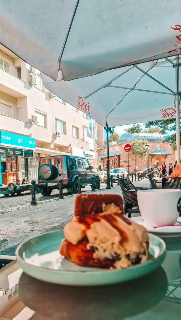 A close-up of a decadent dessert topped with ice cream and caramel sauce, set on an outdoor café table under a Coca-Cola umbrella. The view overlooks a cobblestone street in Plovdiv with parked cars and a historic brick building in the background.