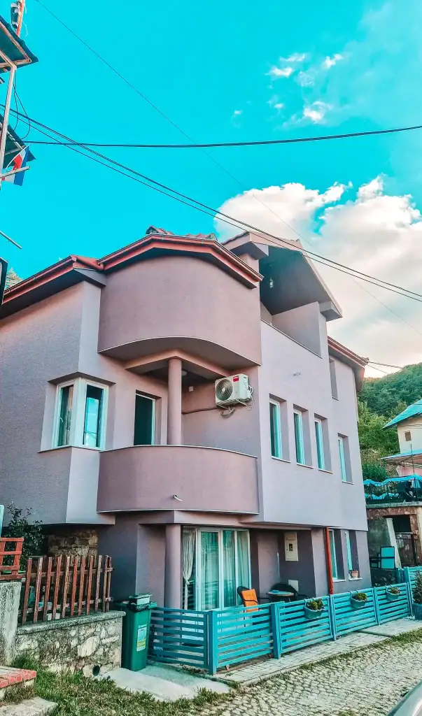 Newly built house with rounded balconies and light purple exterior walls, featuring blue fencing and potted plants along a cobblestone street.