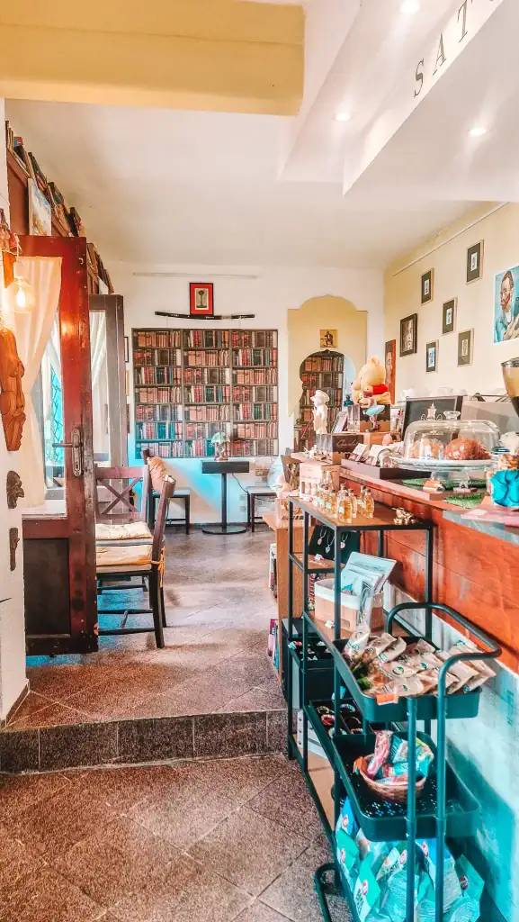 A quaint and eclectic café interior featuring shelves of books, framed art, plush toys, and a counter with cakes and small gift items. The charming decor adds a unique twist to the café experience in Plovdiv.