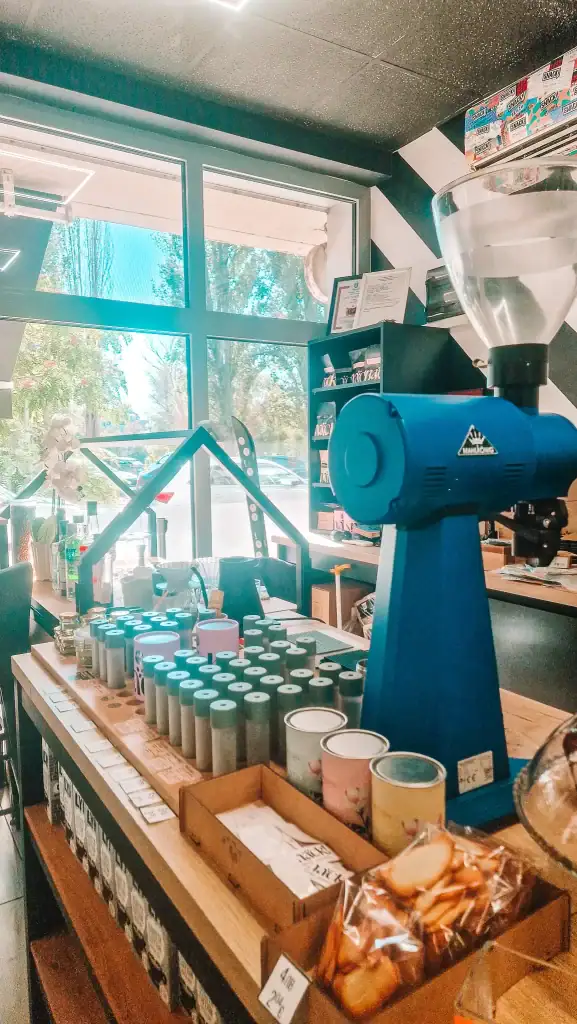 A bright café retail section with a blue grinder, retail shelves, and jars of teas and coffee accessories. Natural light floods in from the large windows, highlighting the café’s modern aesthetic.