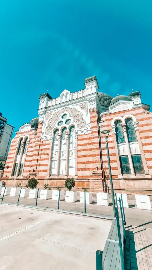Exterior of the Sofia Synagogue, showing striped brickwork, arched windows, and a decorative Moorish façade under a bright blue sky.