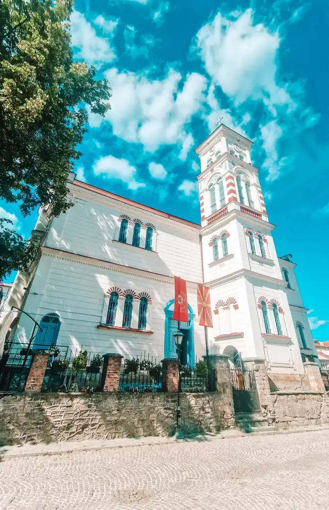 Bright white Orthodox church with red detailing and a tall bell tower, decorated with Macedonian and Krusevo flags, standing against a vivid blue sky.