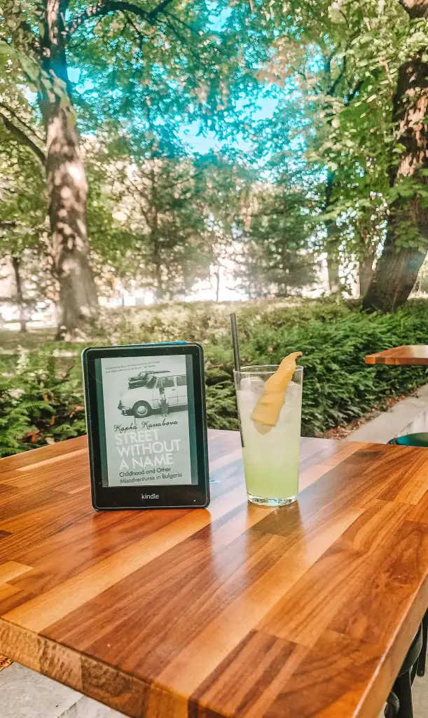 A Kindle displaying Street Without a Name by Kapka Kassabova sits on a wooden café table next to a tall glass of lime soda. The lush greenery and dappled light provide a peaceful setting for reading one of the most insightful books about Bulgaria.