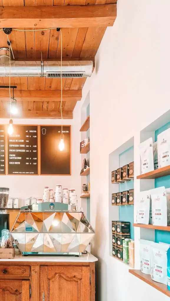 Inside a minimalist café with wooden ceiling beams and a striking mirrored espresso machine. Shelves display artisan coffee bags and the menu lists espresso-based drinks, reflecting the café’s commitment to quality.