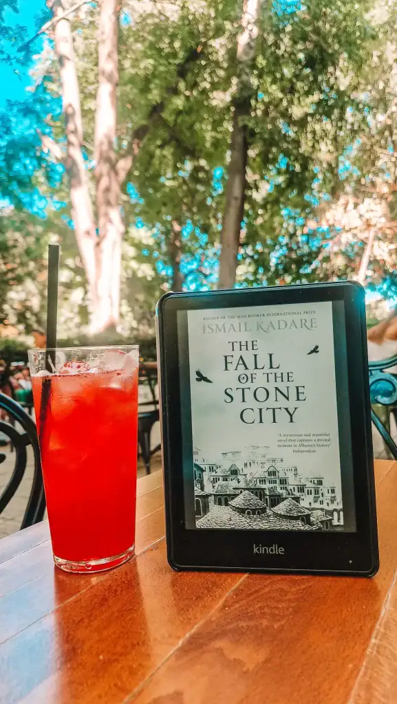 An e-reader displays the cover of The Fall of the Stone City by Ismail Kadare, placed on a wooden table beside a bright red drink. Sunlight filters through tall trees, setting the tone for reading this haunting novel set in Albania during WWII.
