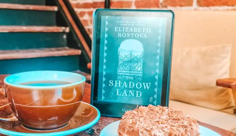 A Kindle open to The Shadow Land by Elizabeth Kostova leans beside a brown cup of coffee and a dessert-topped doughnut. The exposed brick and quiet café ambiance set the tone for diving into one of the best novels set in Bulgaria.