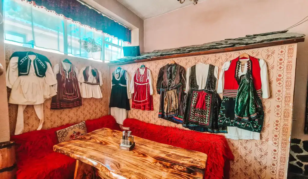 Interior of a cosy room with wooden furniture and red fur-covered benches, featuring a display of traditional Balkan garments hung on the wall, including embroidered vests and dresses in rich fabrics.