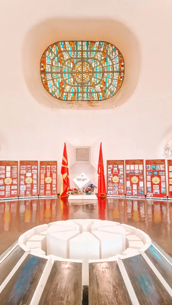 Inside the Makedonium memorial, showing the tomb of revolutionary Nikola Karev flanked by Macedonian flags, flower arrangements, and vibrant red exhibition panels under a stained-glass skylight.
