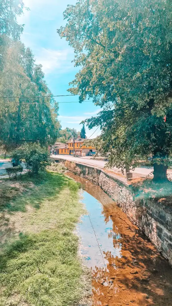 Shallow stream running alongside a grassy path and stone wall, with traditional yellow homes and trees lining the opposite side. A walk along the river offers a quiet contrast to museum hopping.