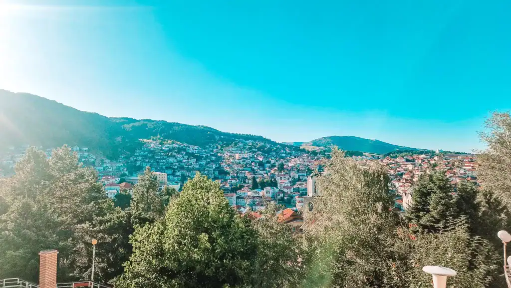 Wide panoramic view of Krusevo’s hillside homes nestled in green forested slopes, taken from an elevated viewpoint under a clear blue sky.