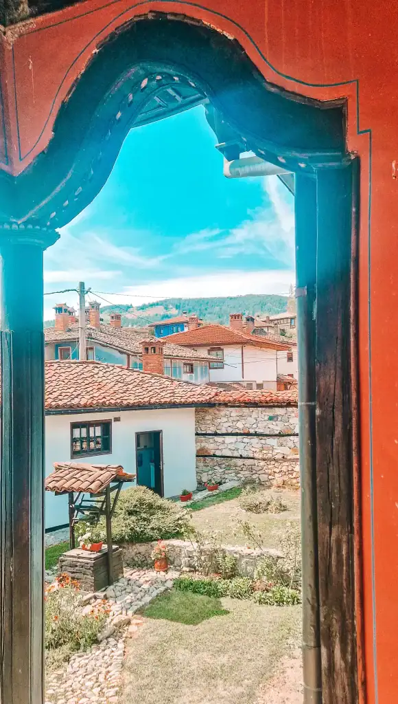 Framed view from under a red-painted archway overlooking a stone courtyard garden with a water well, flower beds, and whitewashed buildings with red tiled roofs. The scene captures the peaceful charm of Koprivshtitsa’s historic homes and gardens.