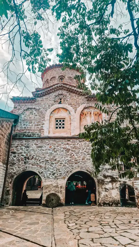 Exterior of Bachkovo Monastery’s stone church, with arched entrances and Byzantine brickwork framed by leafy trees.