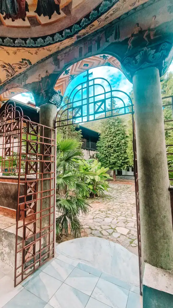 View through the painted archway and iron gates of Bachkovo Monastery, looking onto the courtyard with palm trees and cobbled paths.