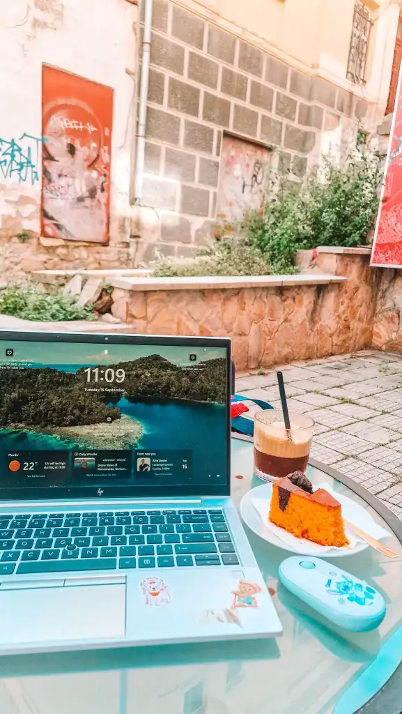 Em works outside at a small café table in Plovdiv with her laptop, an iced coffee, and a slice of vibrant orange cake—making the most of remote work in a sunlit urban setting.