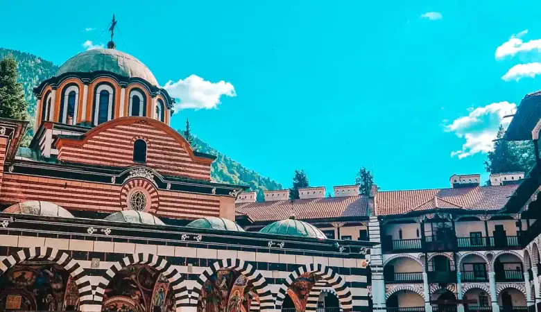 Striking striped arches and colourful frescoes adorn the church facade in the courtyard of Rila Monastery, with forested mountains in the background — one of the most popular day trips from Sofia.