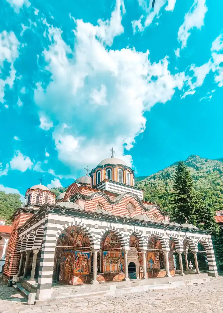 Full façade of Rila Monastery with frescoed arches and domed roof beneath a bright blue sky.