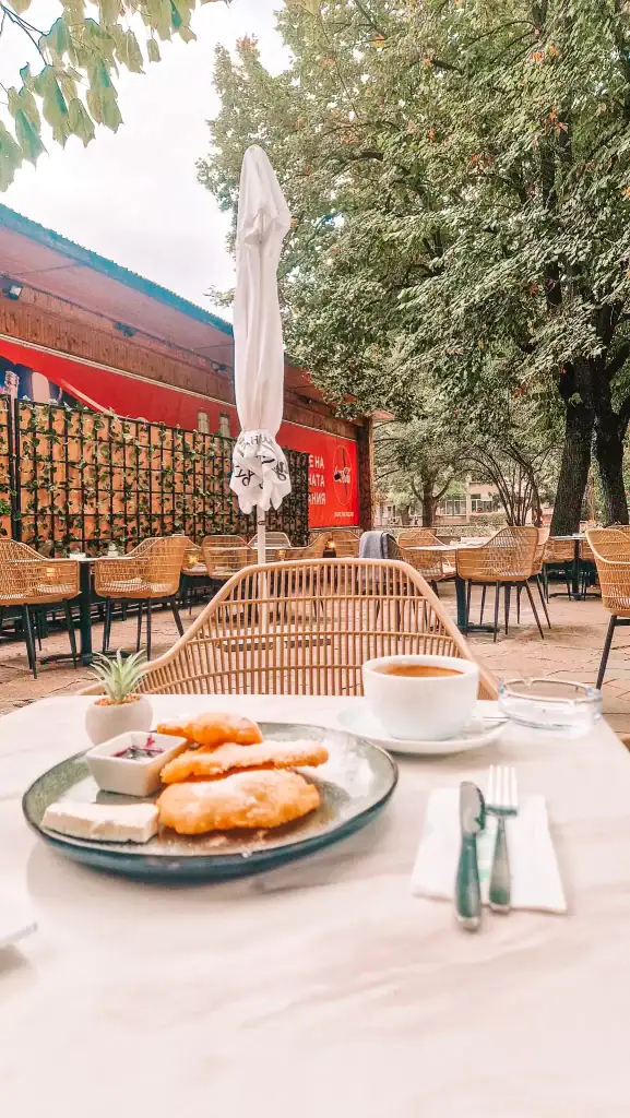 A shaded café garden in Plovdiv with a plate of Bulgarian mekitsi, jam, and white cheese next to a fresh cup of coffee—great for a remote work morning in the fresh air.